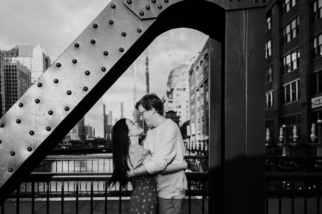 Black and white photo of engaged couple holding each other as they stand on the Clark Street Bridge in downtown Chicago during sunrise engagement session