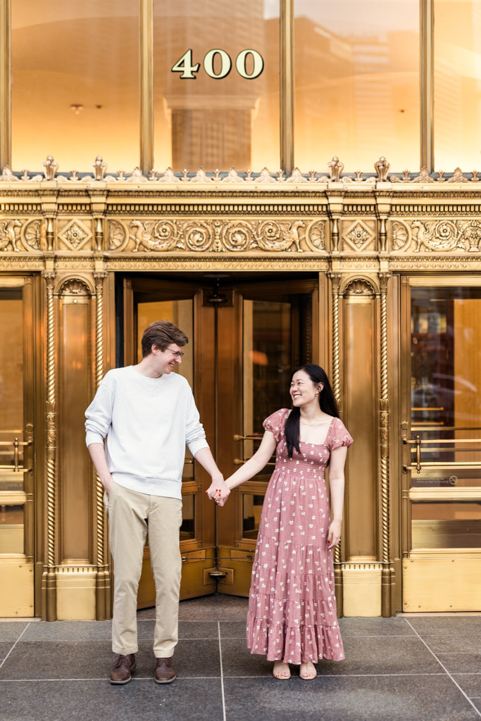 Engaged couple holds hands in front of golden doors at the Wrigley Building in downtown Chicago