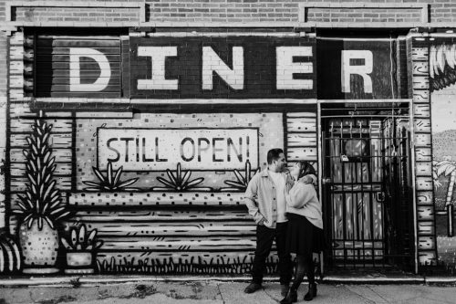 Black and white photo of couple smiling at each other in front of a mural with "Diner Still Open" text in Humboldt park Chicago