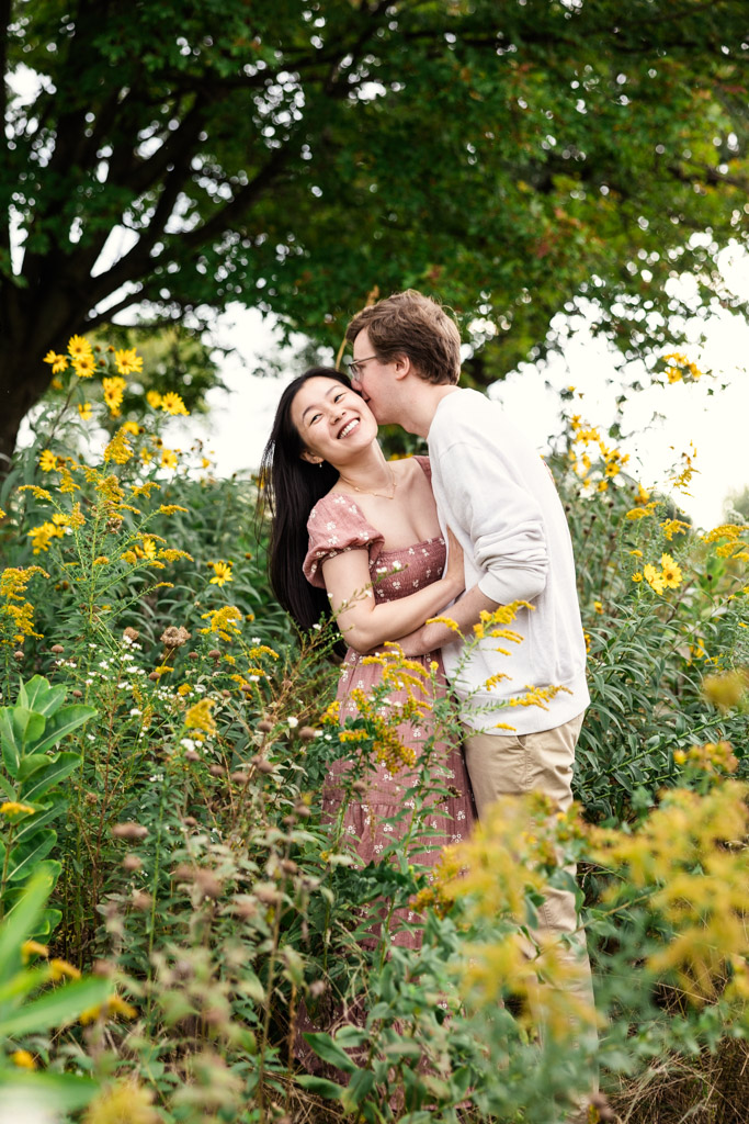 Man kisses his partner's cheek as they stand in a field of yellow wildflowers in Lincoln Park during sunrise engagement session