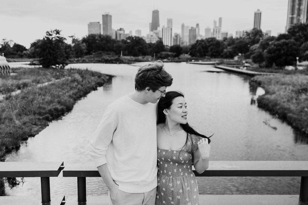 Black and white photo of engaged couple on a Lincoln Park bridge with a pond and downtown Chicago skyline in the background