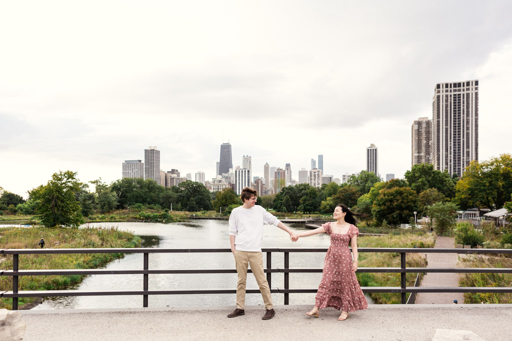 Man leads his partner across a bridge in Lincoln Park with downtown Chicago skyline in the background during sunrise engagement session