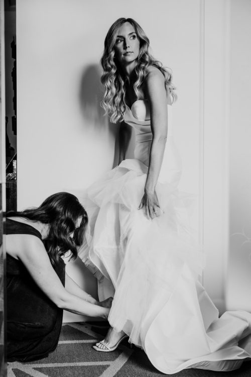 Black and white photo of bride in a wedding dress getting help with her shoes