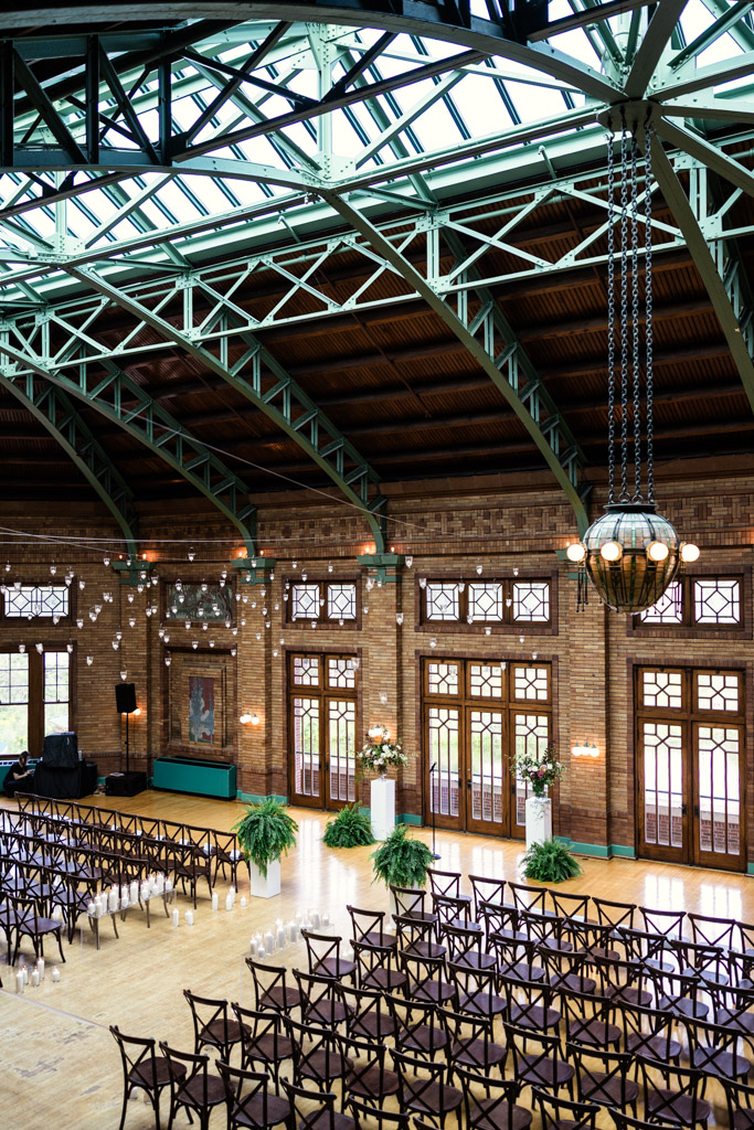 Cafe Brauer wedding ceremony space features rows of chairs on a polished wooden floor, large arched windows, and metal-framed glass ceiling