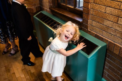 Happy child in a white dress leans on a teal radiator during wedding reception at Cafe Brauer in Lincoln Park Chicago