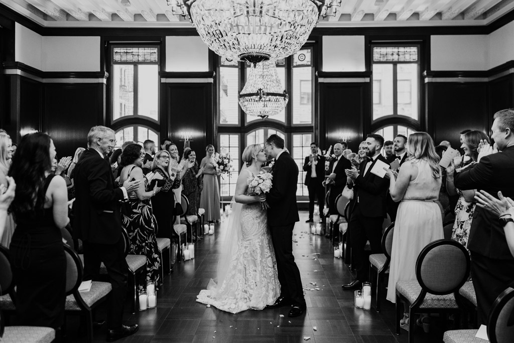 Bride and groom kiss while guests applaud during their Chicago Athletic Association Hotel wedding ceremony in the Madison Ballroom