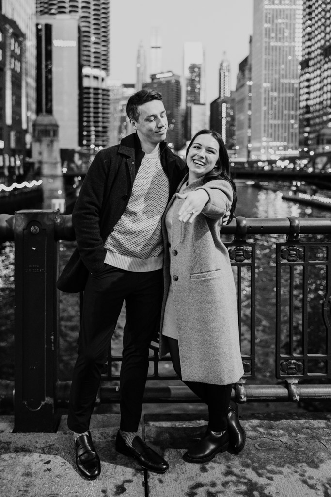 Black and white photo of happy couple standing on a downtown Chicago bridge, the woman proudly showing off her ring during fall engagement session
