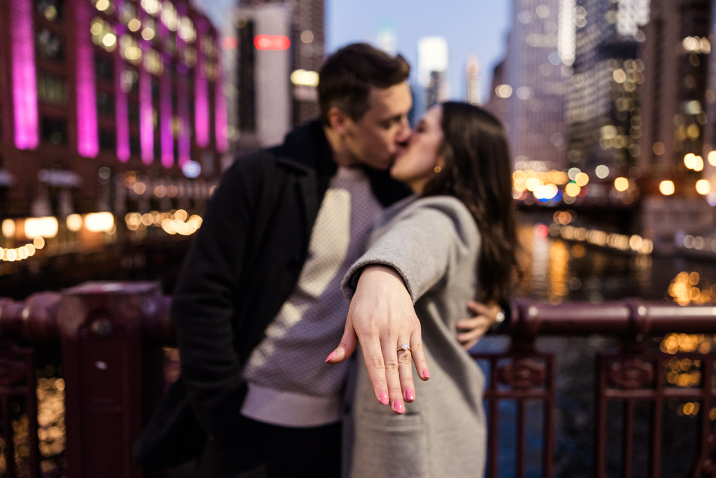 Creative photo of engaged couple kisses on a downtown Chicago bridge with focus on the woman's outstretched hand and her engagement ring and city lights in the background
