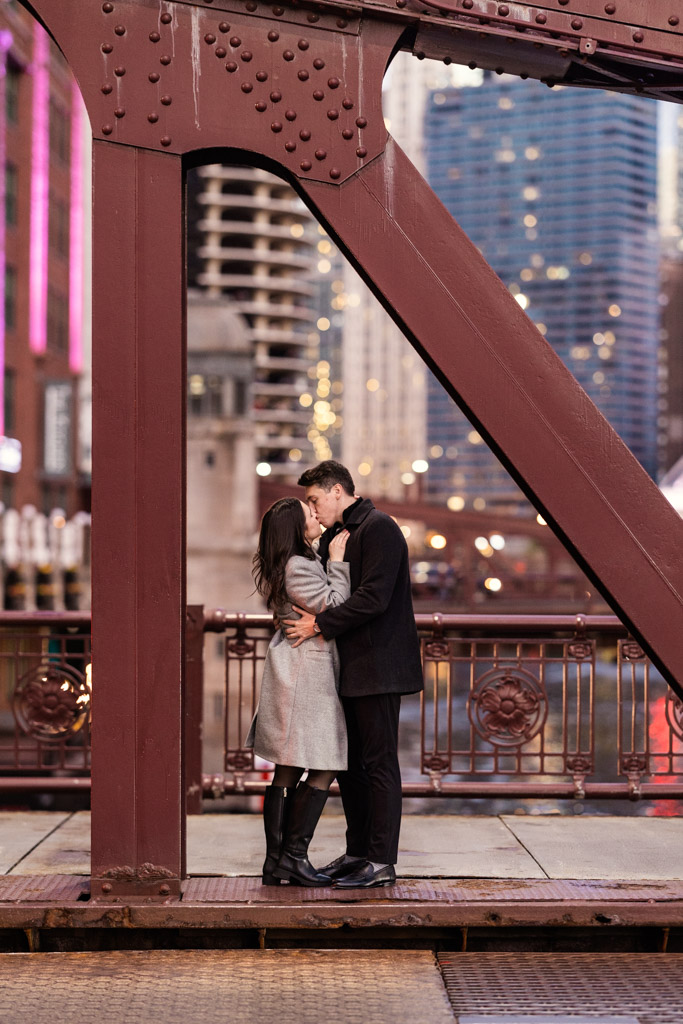 Couple kisses on the LaSalle Street Bridge in downtown Chicago with city lights and buildings in the background during their fall engagement session