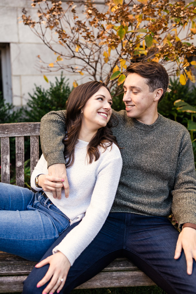 Happy couple sits on a bench with vibrant autumn leaves in the background during Lincoln Park engagement session