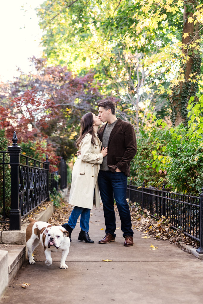 Couple kisses on a Lincoln Park path with their dog walking nearby during fall engagement session
