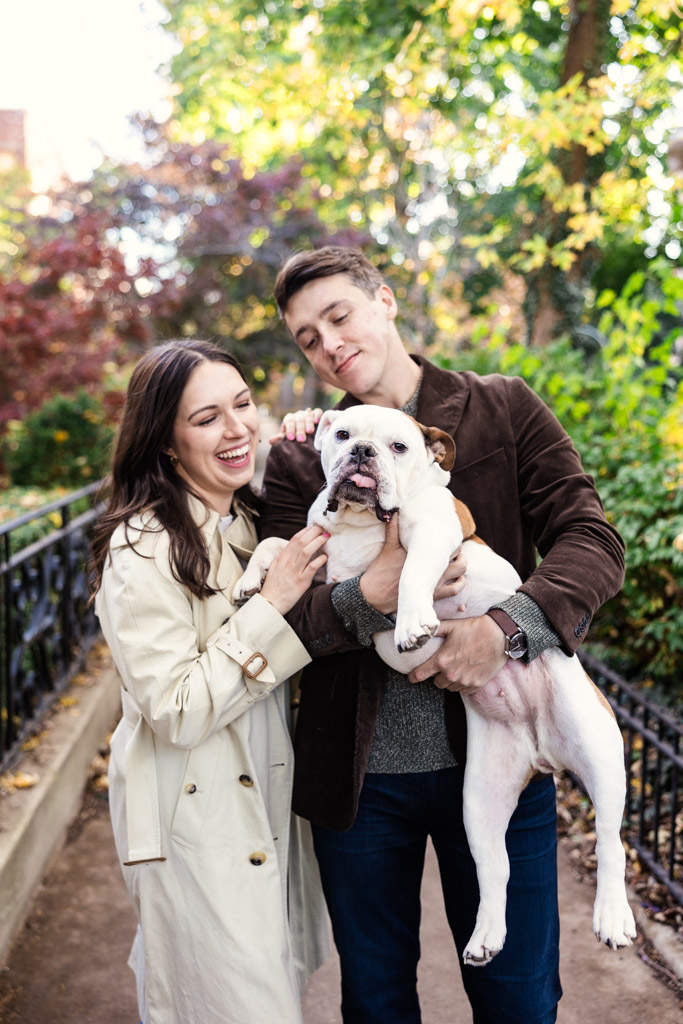 Engaged couple holds their bulldog in Lincoln Park, surrounded by lush greenery and vibrant autumn colors