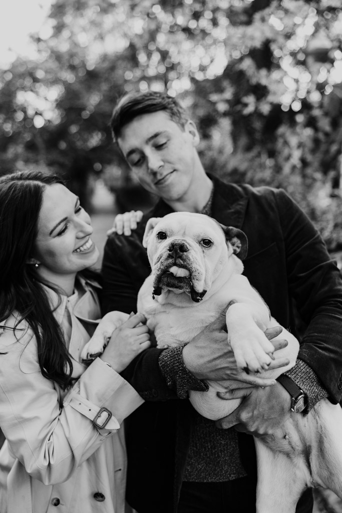 Happy couple smiles and holds a bulldog outdoors during their fall engagement session