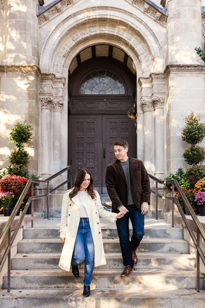 Couple walks down steps holding hands, framed by a large arched doorway during fall Lincoln Park engagement session
