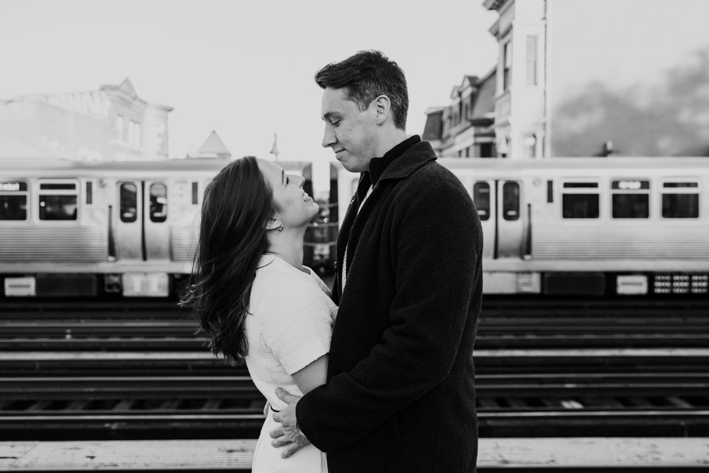 Black and white photo of couple embracing on a CTA platform with a train in the background during fall engagement session