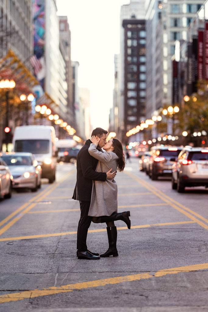 Couple kisses on the State Street median in downtown Chicago with cars and tall buildings in the background during fall engagement session
