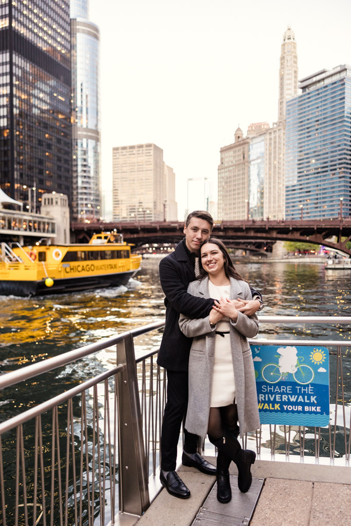 Portrait of engaged couple standing on the edge of the Chicago River with downtown skyscrapers and a yellow water taxi in the background