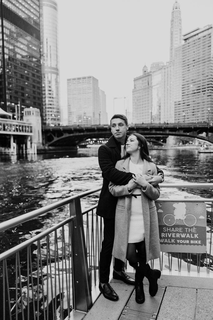 Black and white photo of engaged couple standing on the Chicago Riverwalk with tall buildings and a bridge in the background