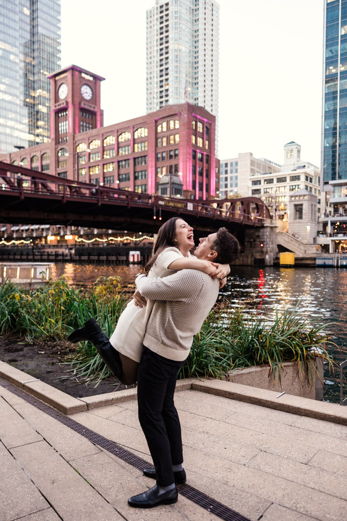 Man lifts his partner as they stand on the Chicago Riverwalk near the LaSalle Street Bridge during fall engagement session
