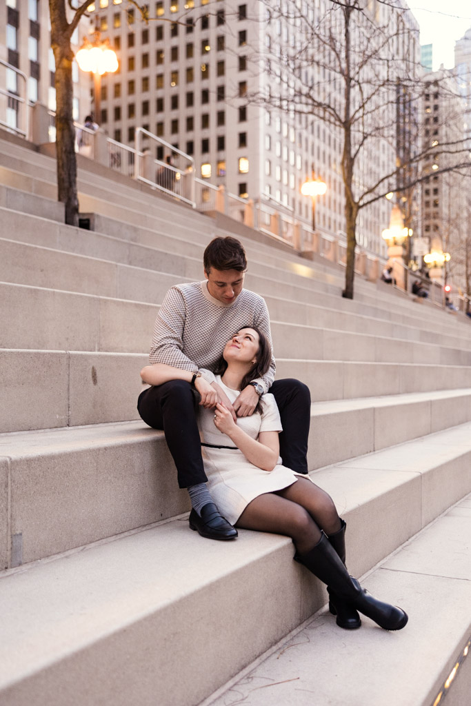 Couple sits on outdoor steps at the Chicago Riverwalk with downtown skyscrapers in the background during fall engagement sesson