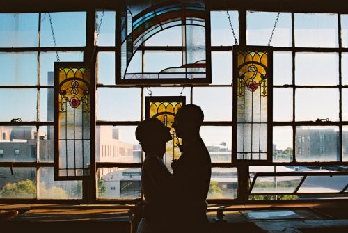 Silhouetted bride and groom stand by vintage stained glass windows in Salvage One, with Chicago city buildings as their backdrop