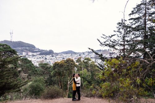 Engaged couple kisses on Strawberry Hill with San Francisco cityscape and Sutro Tower in the background