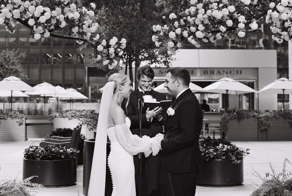 Under a floral arch, the bride and groom exchange vows with the officiant nearby, all set against the breathtaking backdrop of South Branch Tavern and Grille's outdoor setting.