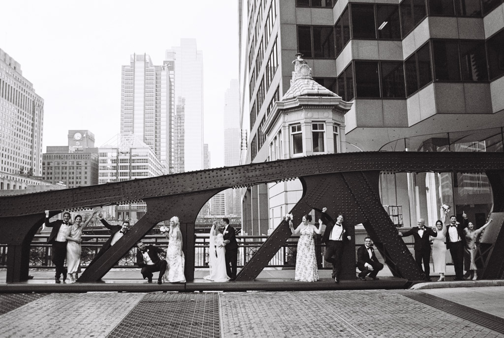 A wedding party poses elegantly on a bridge with a city skyline in the background, captured in timeless black and white. The scene evokes the charm of a celebration at South Branch Tavern and Grille, blending urban sophistication with heartfelt joy.