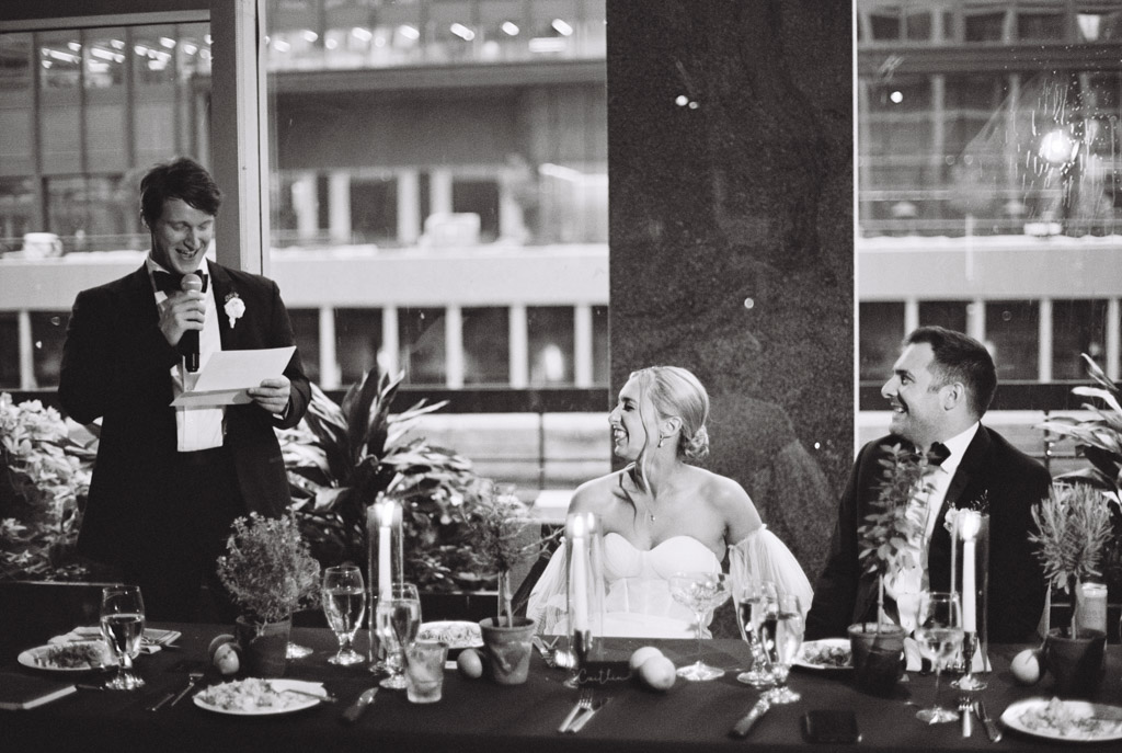 A man gives a heartfelt speech at a wedding reception in South Branch Tavern and Grille as the bride and groom smile, seated at their beautifully decorated table. Black and white photo.