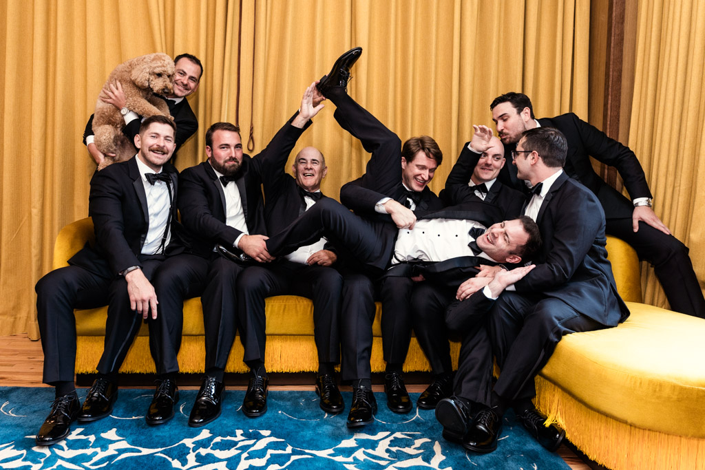Fun photo of groom and groomsmen sitting on a yellow couch before his South Branch Tavern and Grille weddiing, playfully posing with a dog in front of yellow curtains