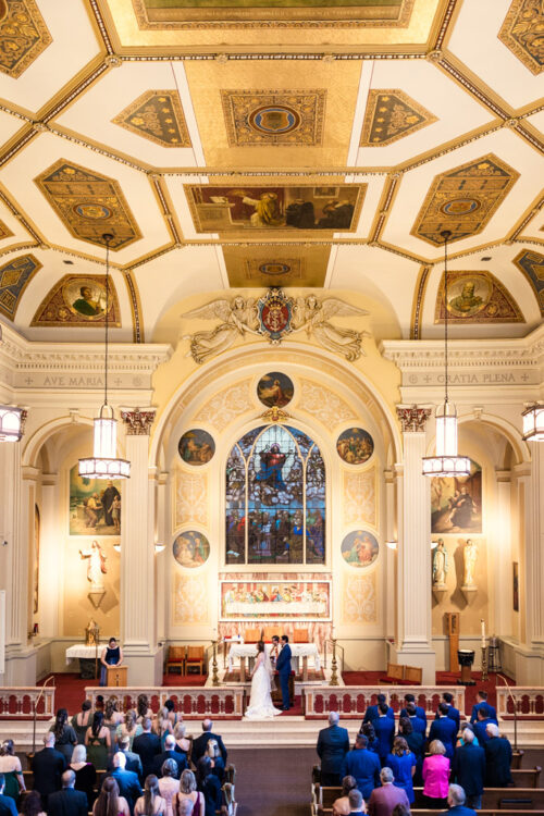 Bride and groom stand at the altar of Assumption Catholic Church surrounded by guests