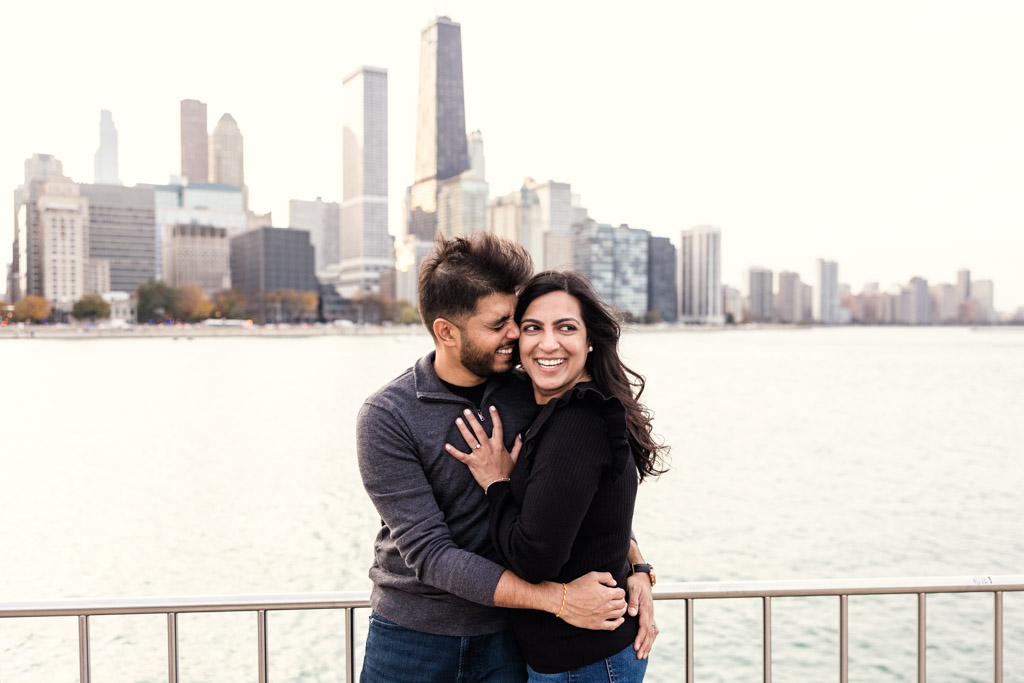 A couple embraces and smiles on the Olive Park lakefront with Chicago skyline in the background, during fall engagement session