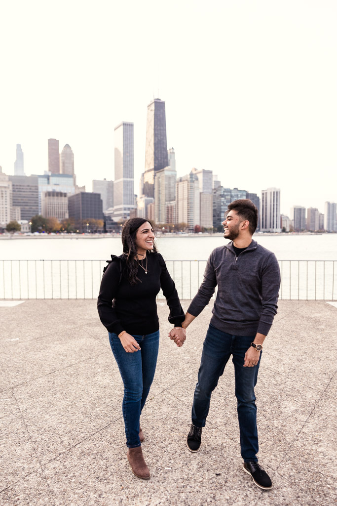 A couple holds hands and smiles as they walk along the lakefront in Olive Park with the Chicago skyline in the background