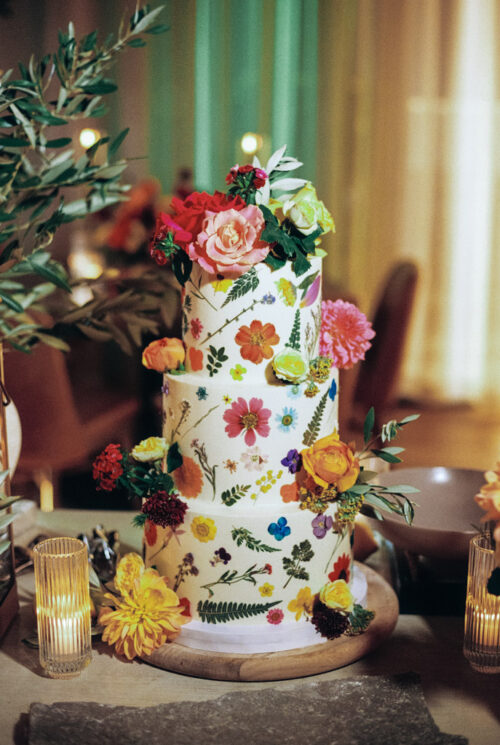 A four-tiered wedding cake adorned with colorful edible flowers and leaves, reminiscent of a film photography snapshot, is beautifully surrounded by flickering candles.