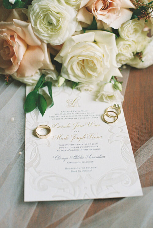 Wedding invitation with gold rings, elegantly captured through film photography, surrounded by white and peach roses on a sheer fabric.