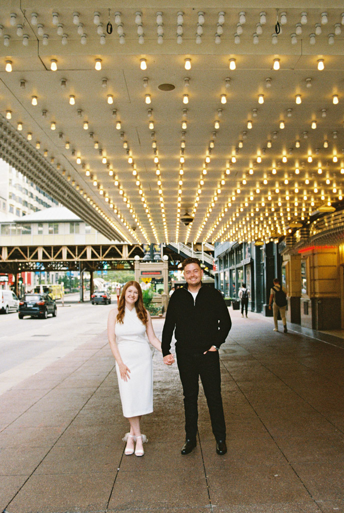 A couple holding hands under a ceiling of lights on a city street, their moment captured with the nostalgic charm of film photography.