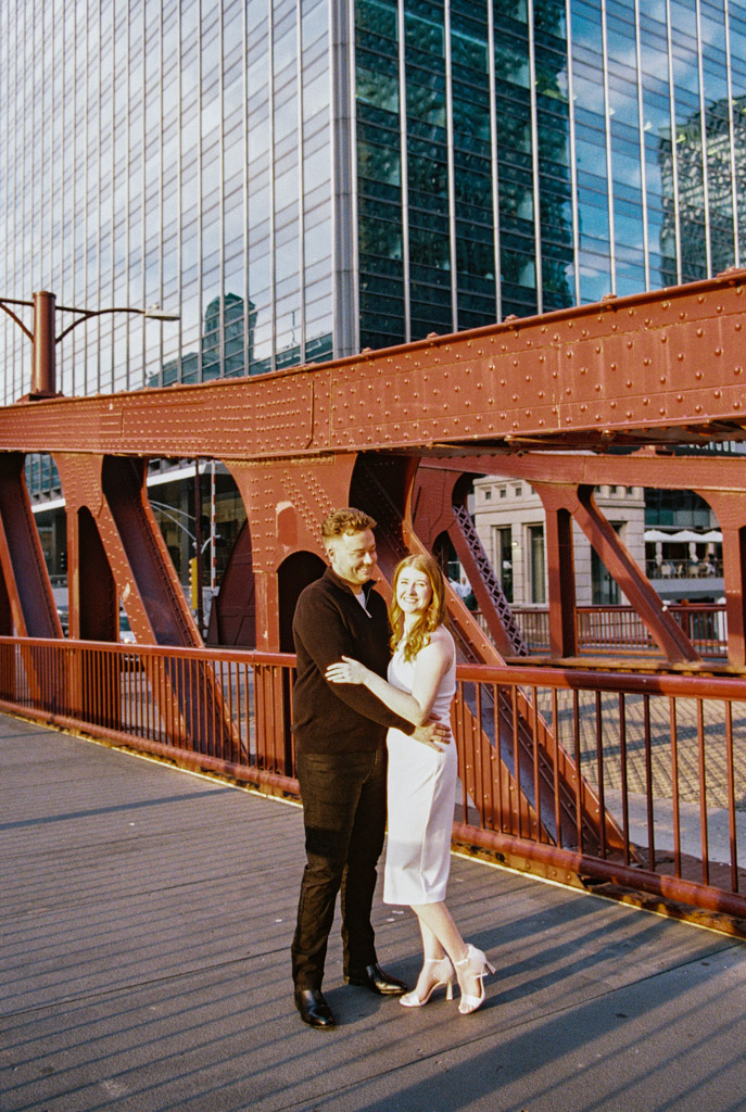 A couple embraces on a red bridge against a backdrop of modern glass buildings, their moment beautifully captured through the nostalgic lens of film photography.