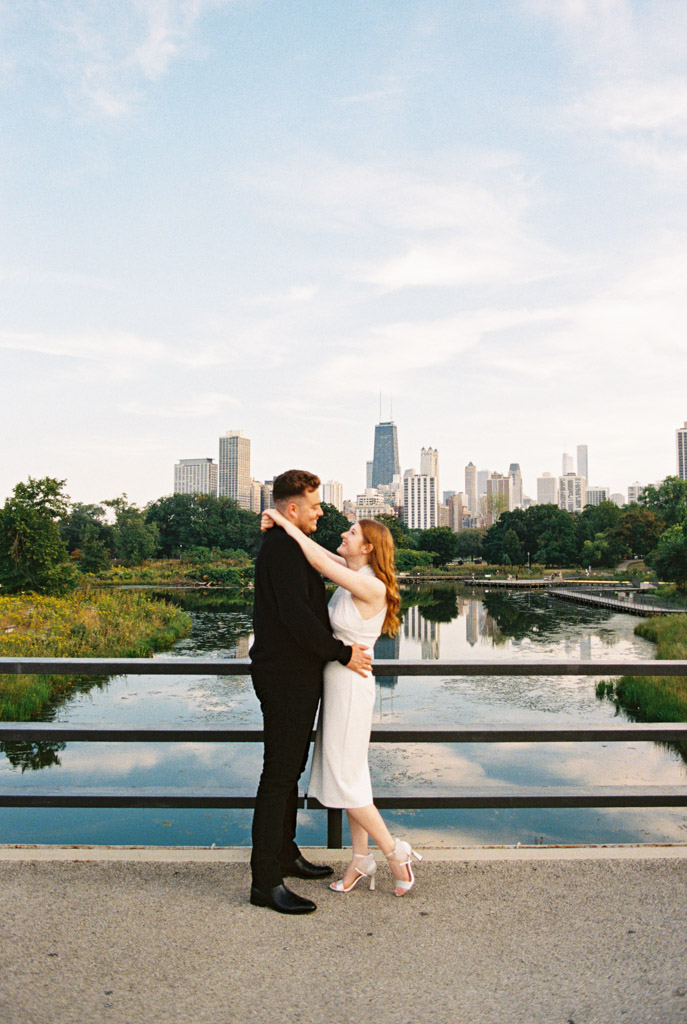 A couple embraces on a bridge with a city skyline and lake in the background under a cloudy blue sky, their moment captured through the nostalgic lens of film photography.