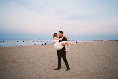 Man carries his partner across the sand at North Avenue Beach at sunset during downtown Chicago engagement session
