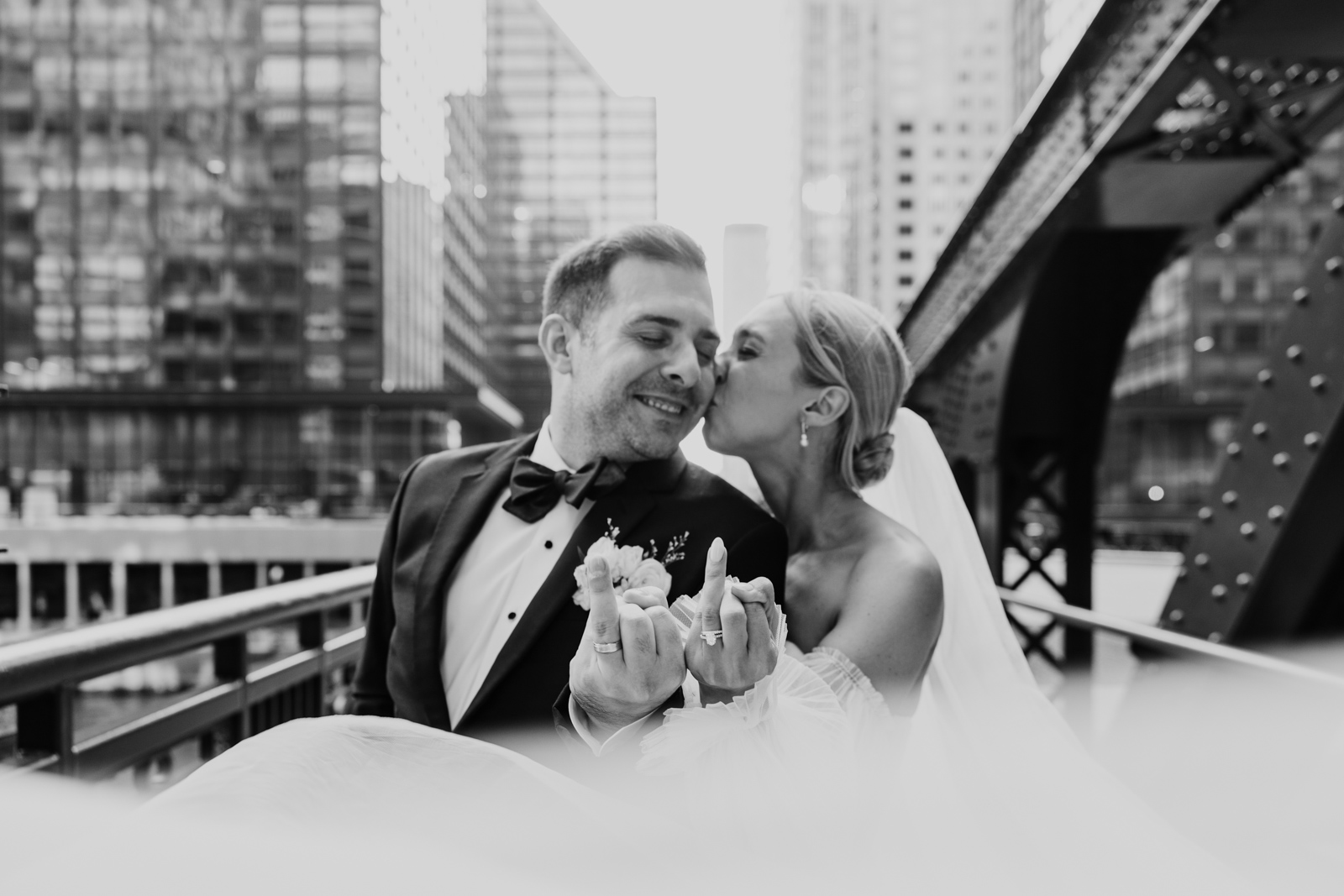 Bride kisses groom Monroe Street Bridge while showing off wedding rings with veil floating in foreground