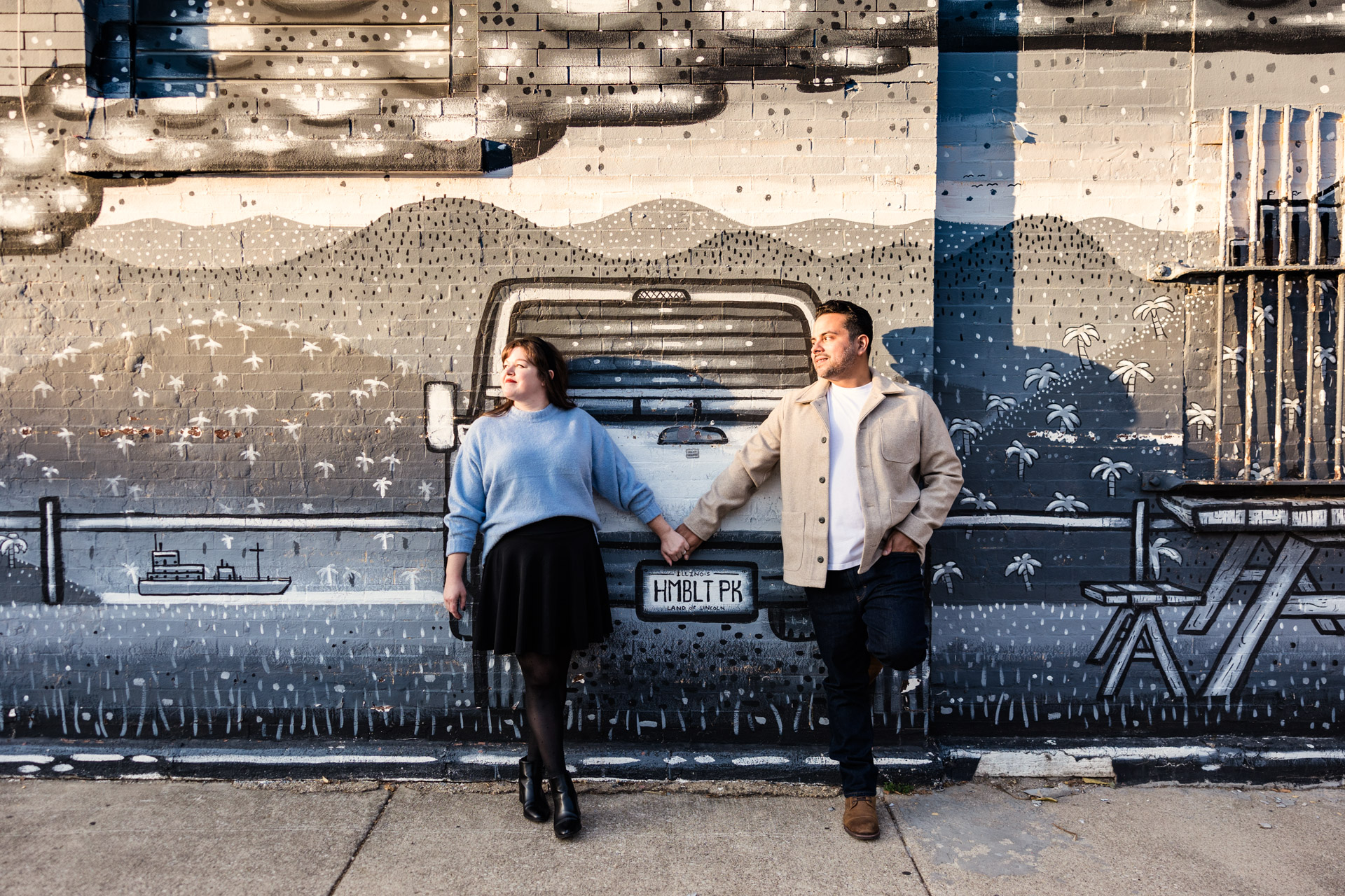 A couple holds hands in front of a grayscale Sick Fisher mural in Humboldt Park during their neighborhood engagement session