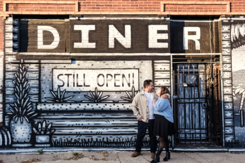 Photo of couple smiling at each other in front of a mural with "Diner Still Open" text in Humboldt Park Chicago