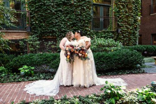 Brides in elegant dresses kiss, holding vibrant bouquets in the courtyard at Ivy Room Chicago