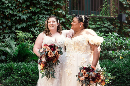 Two brides in white dresses, holding bouquets, standing together in the courtyard at Ivy Room Chicago