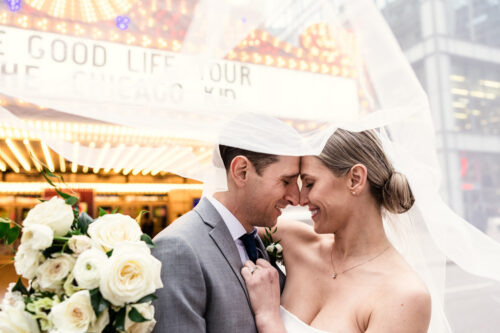 A bride and groom smile under a veil, holding a bouquet of white roses in front of a lit marquee sign, capturing the best of 2024.