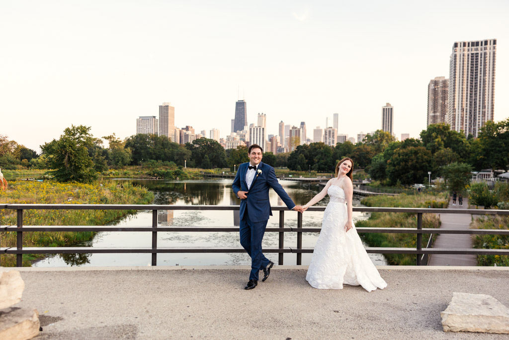 Bride and groom pose holding hands on a bridge in Lincoln Park before their wedding with downtown Chicago in the background