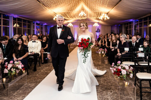 A bride walks down the aisle with an older man in a decorated indoor venue, surrounded by seated guests.
