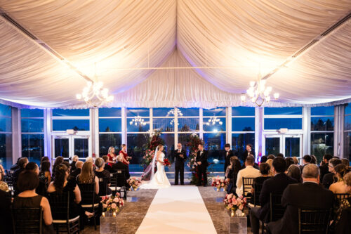 Indoor wedding ceremony with a white aisle, floral decorations, and guests seated under a draped ceiling with chandeliers.
