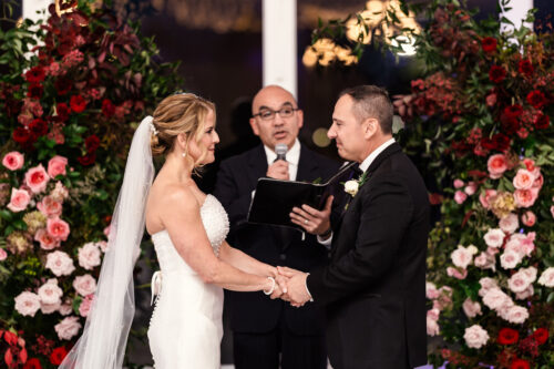 A couple exchanges vows at their wedding, surrounded by vibrant floral arrangements, with an officiant nearby.