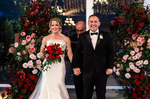 A bride and groom holding hands, standing under a floral arch with red and pink flowers.